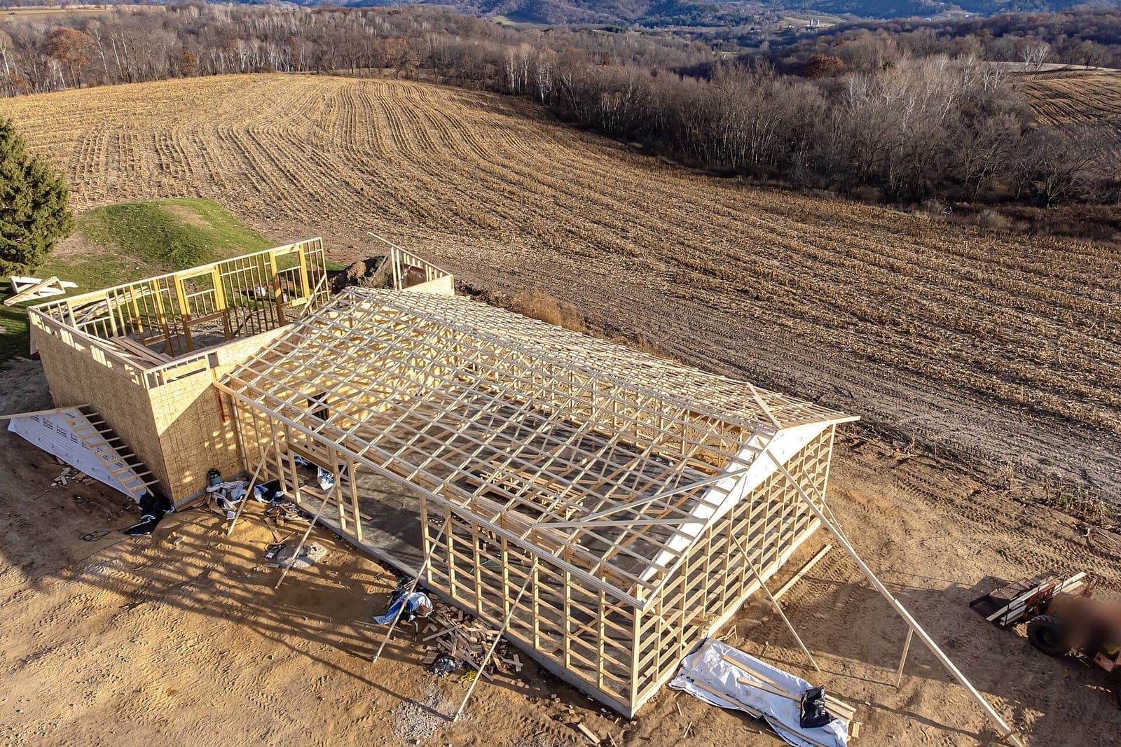 Aerial view of a new wood-framed house and garage with open roof trusses beside a harvested field