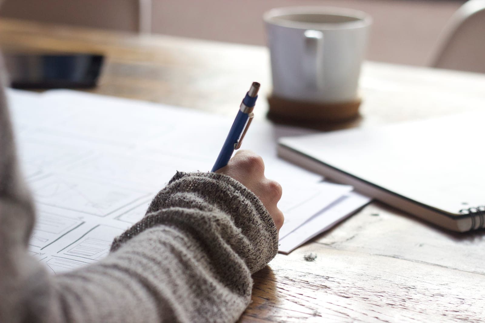 A person sketching app screens on paper at a wooden table next to a coffee cup