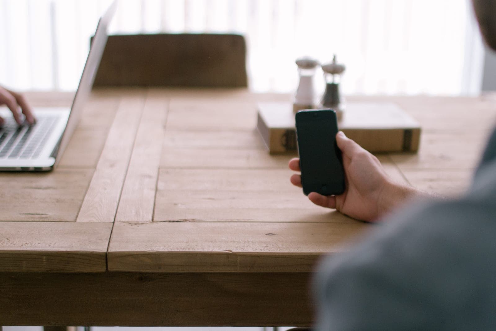 A hand holds a smartphone at a wooden table beside an open laptop