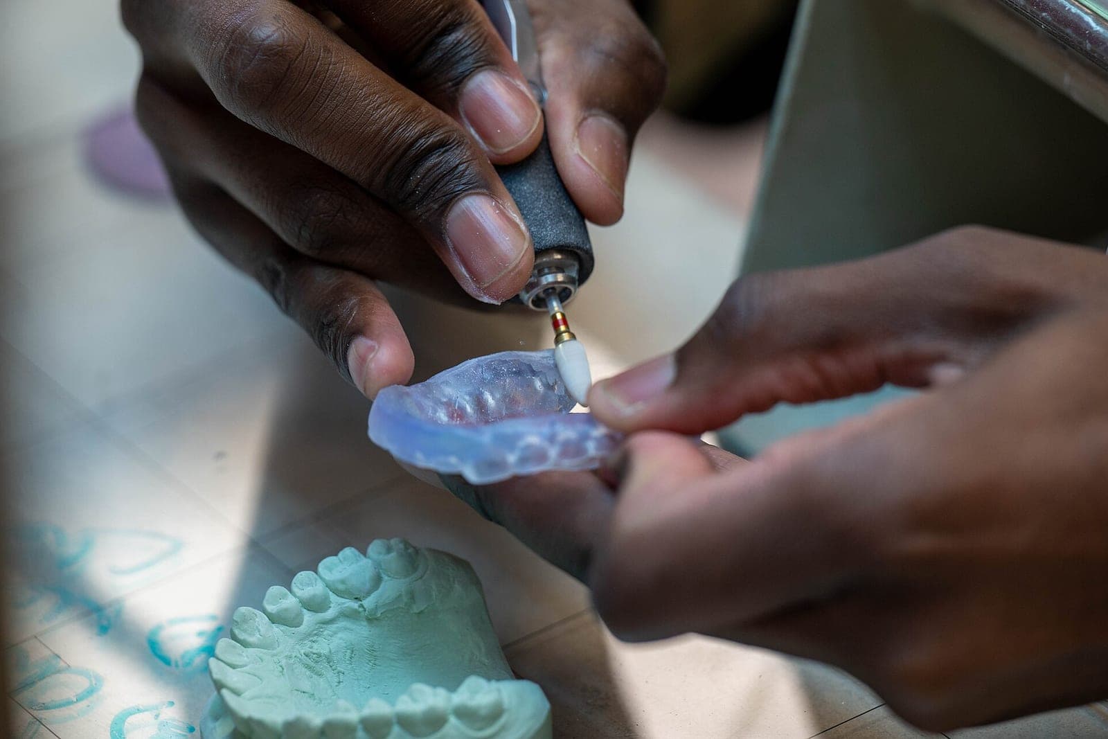 Hands polish a clear mouth guard with a small rotary tool above a pale green stone model of teeth
