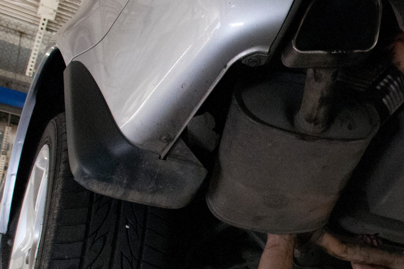 The rear wheel, mud flap, muffler and exhaust tip of a silver car raised on a shop lift, with a mechanic's hands reaching up underneath