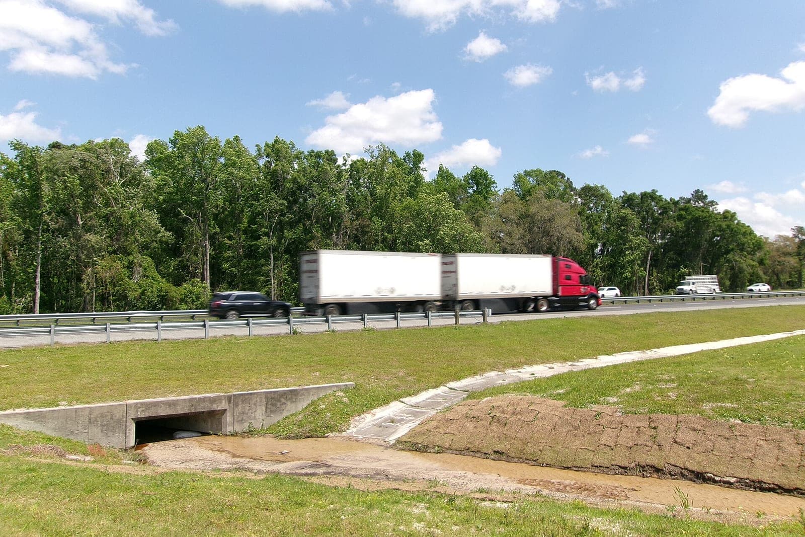 A red tractor pulling two white trailers passes on a divided highway lined with green trees