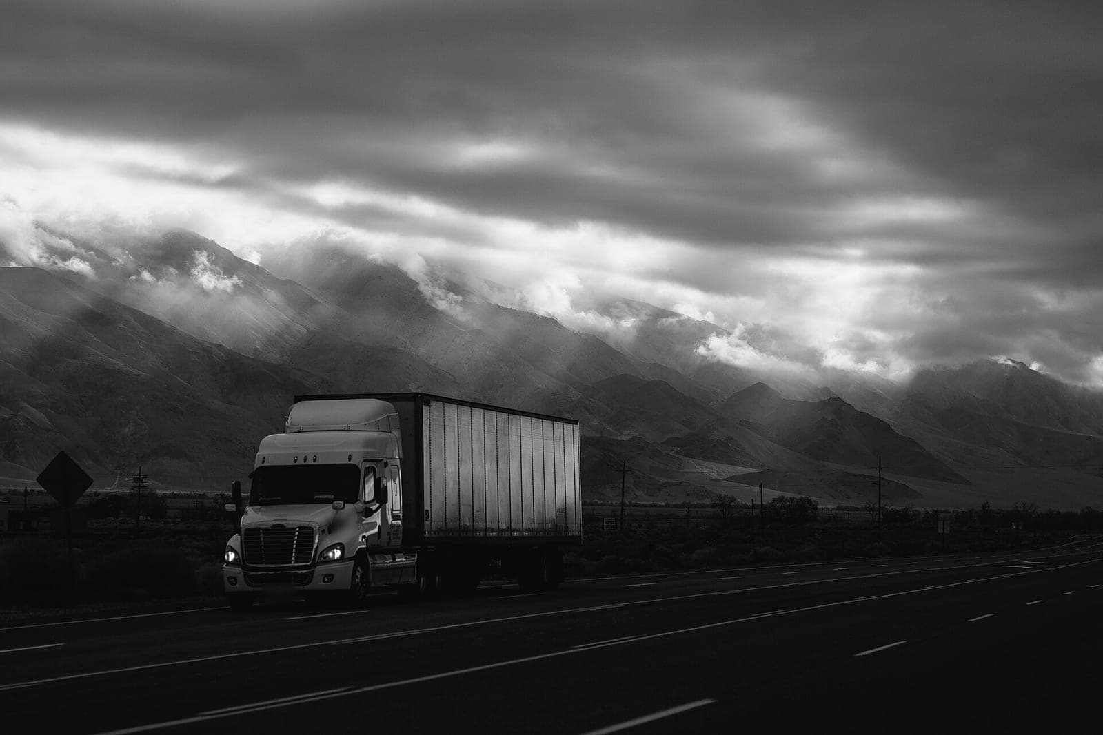 A white semi truck with a box trailer drives along a highway below cloud-covered mountains, shown in black and white