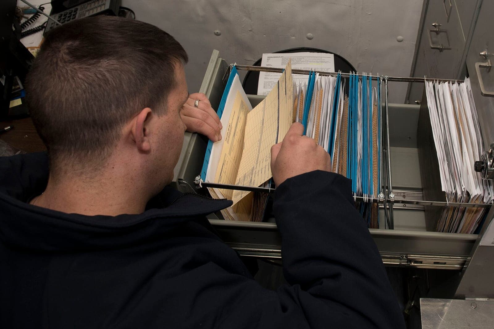 A man seen from behind flips through hanging file folders in an open cabinet drawer