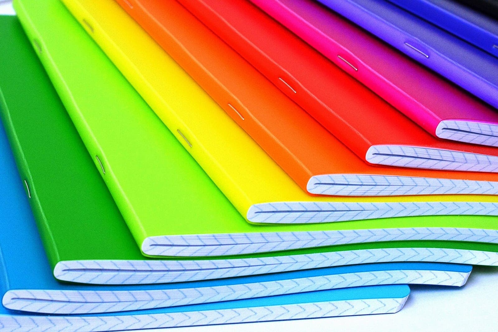 A fanned stack of stapled paper notebooks with bright covers in every color of the rainbow