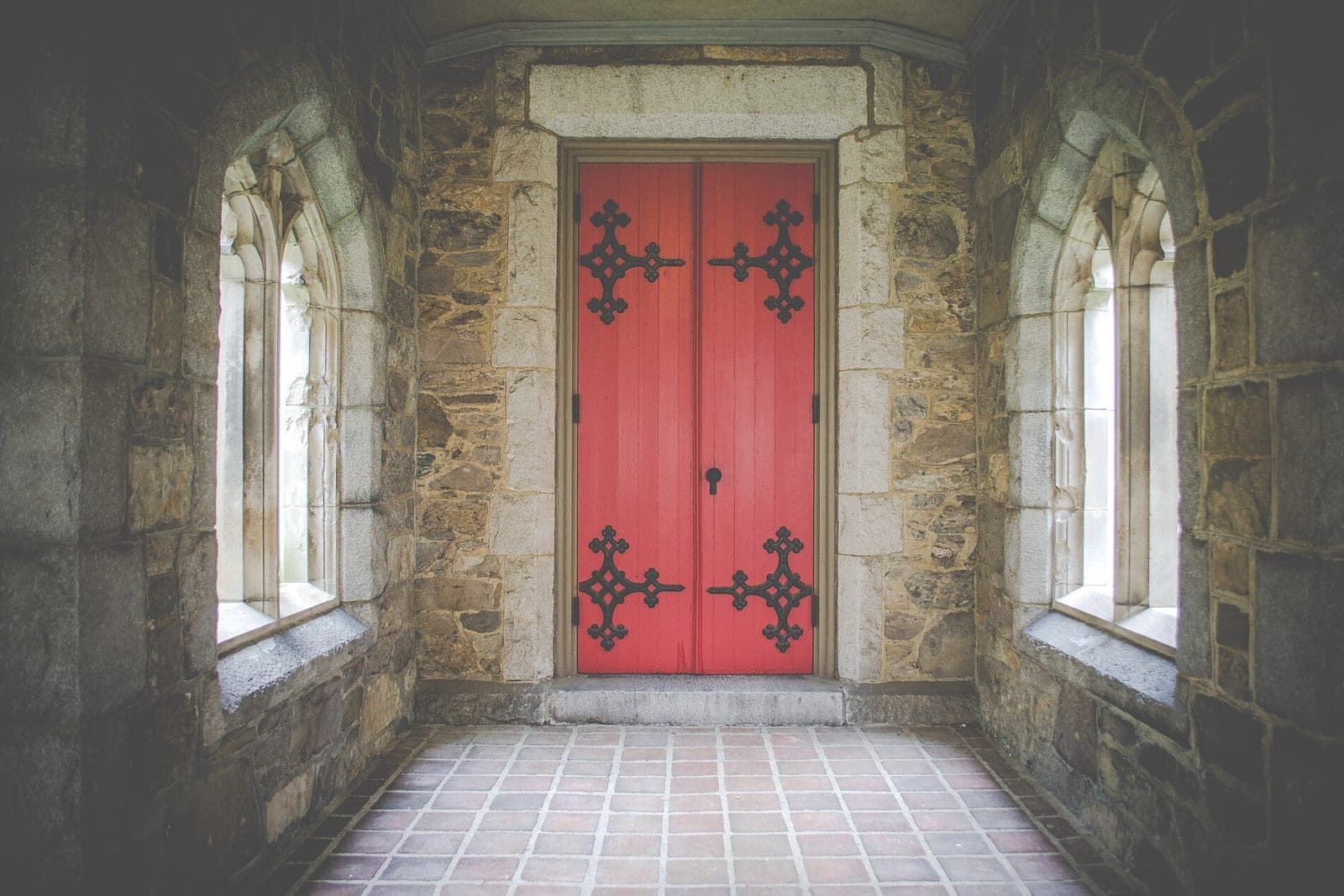 A red double door with black iron hinges at the end of a stone church porch lit by two arched windows