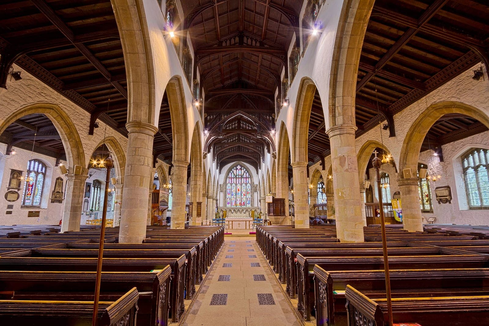 Rows of empty dark wooden pews line the stone-arched nave of a church, with a stained glass window above the altar