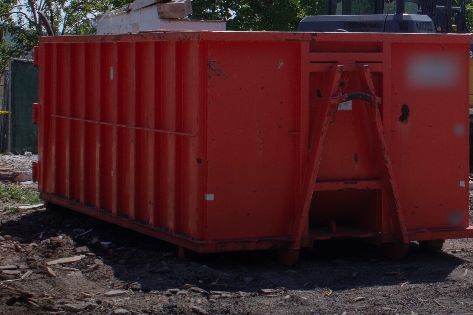 A red roll-off container with its hook bar rests on open dirt at a cleared building lot