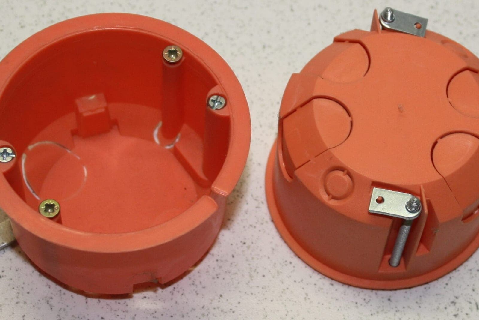 Two orange round electrical boxes with mounting screws on a speckled white counter