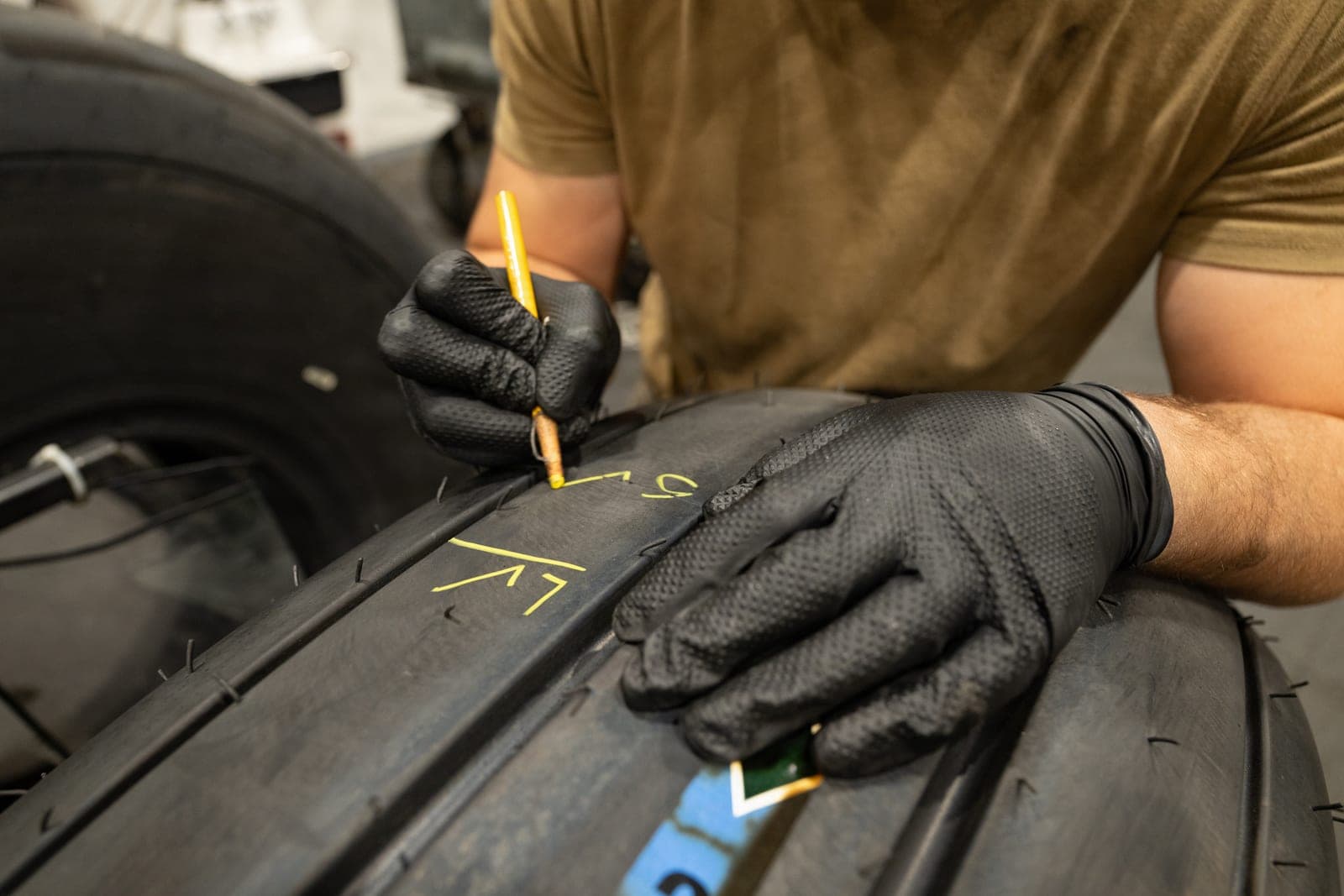 Gloved hands mark a spot on a large tire with a yellow wax crayon in a maintenance shop