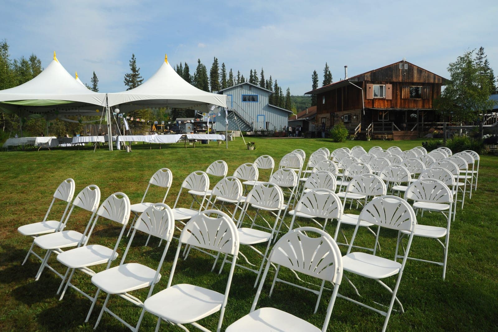 Rows of white folding chairs on a lawn beside two white party tents, with a wooden lodge behind