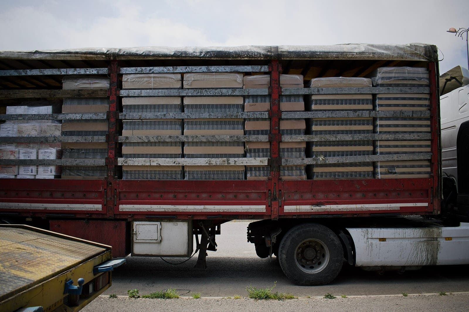 A red truck trailer loaded with stacked, plastic-wrapped cartons behind open side slats, parked in daylight