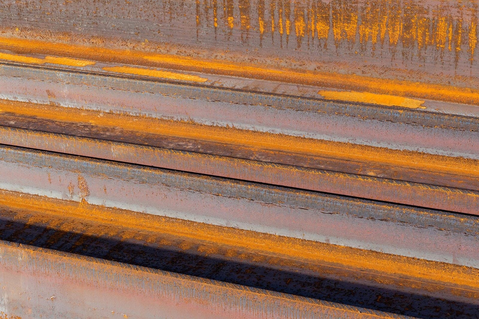 Long steel beams with rusty orange flanges lie stacked side by side in low sunlight