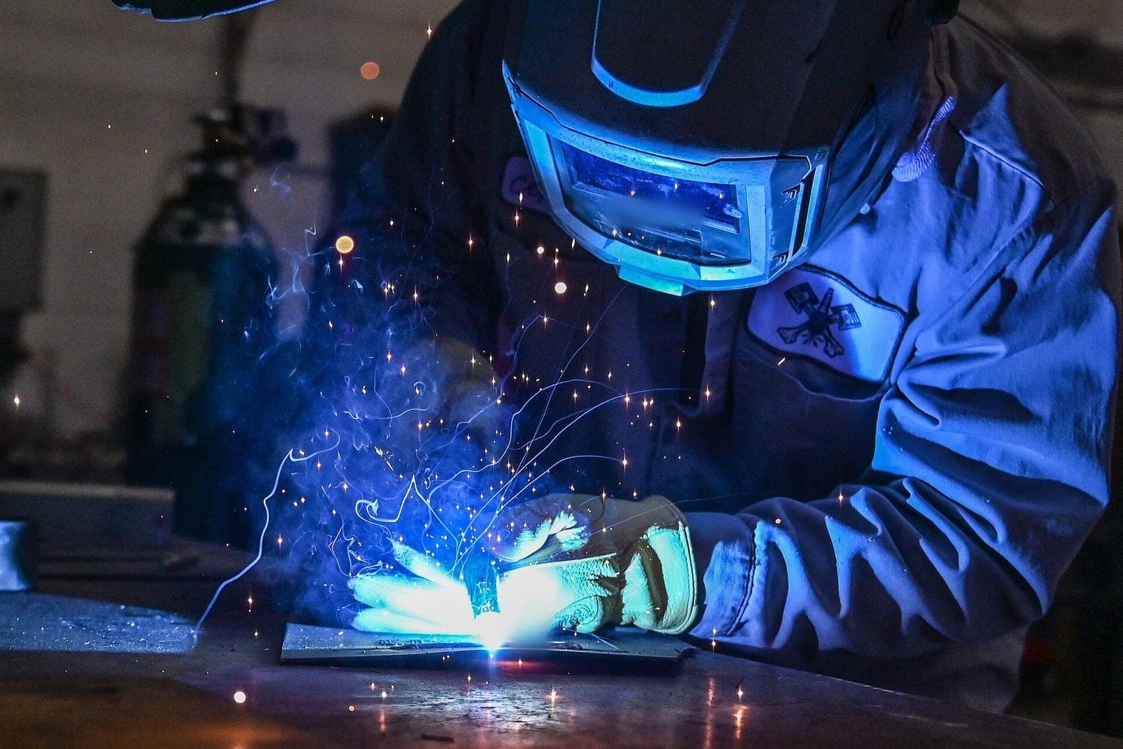 A welder in a dark helmet and leather gloves joins two steel plates as blue sparks scatter across the table