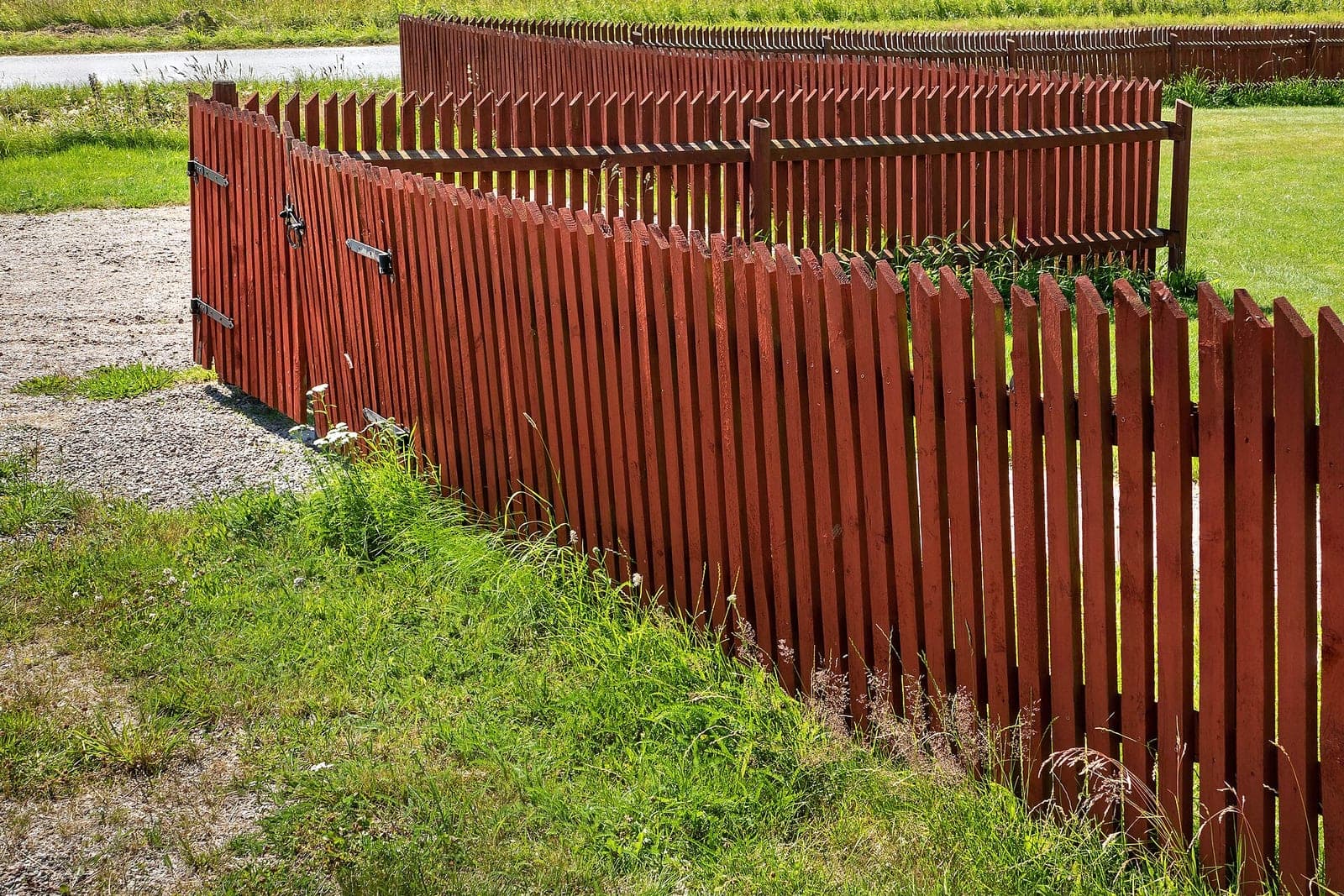 A red wooden picket fence with a gate on black hinges runs along a gravel path and green lawn