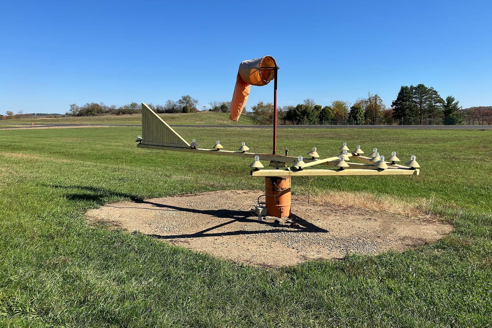 An orange windsock above a wind tee on the grass beside an airport runway on a clear day
