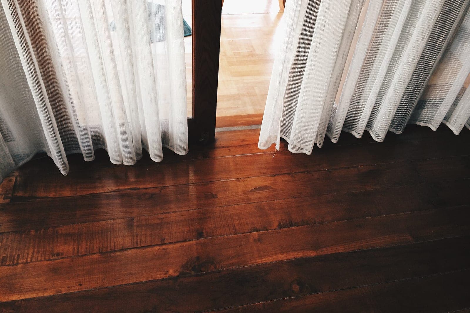 Sheer white curtains hanging over a dark wood plank floor beside an open doorway