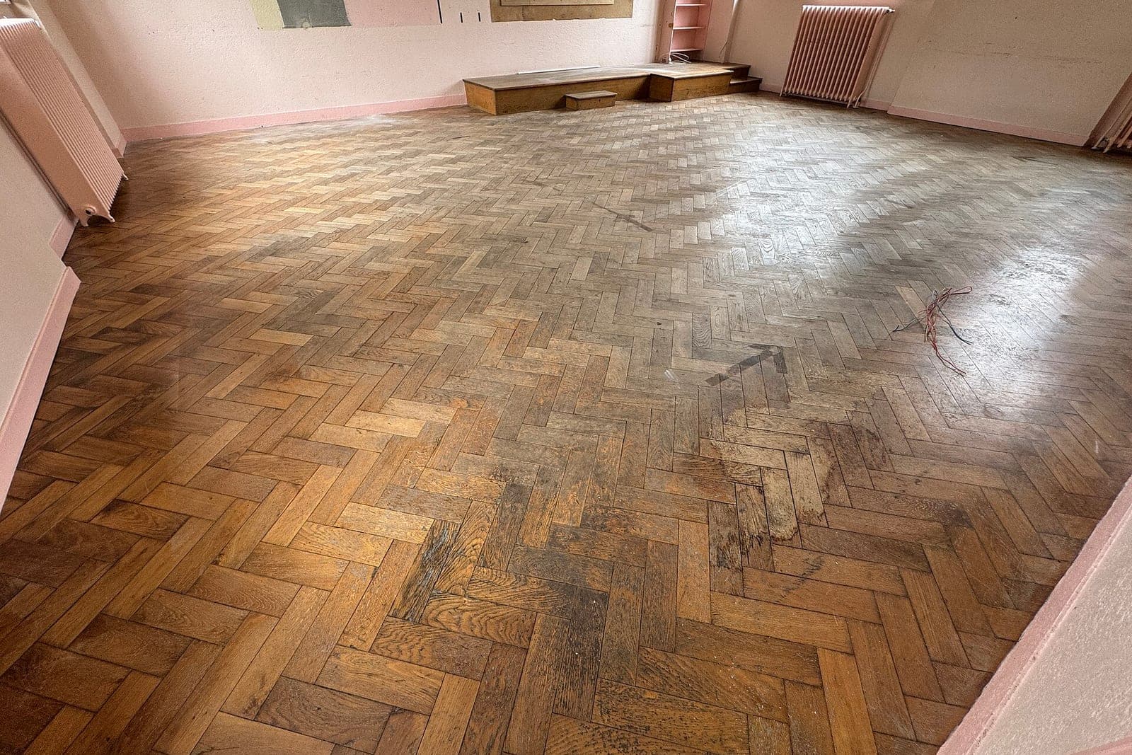 A worn herringbone wood floor in an empty room with pale pink walls