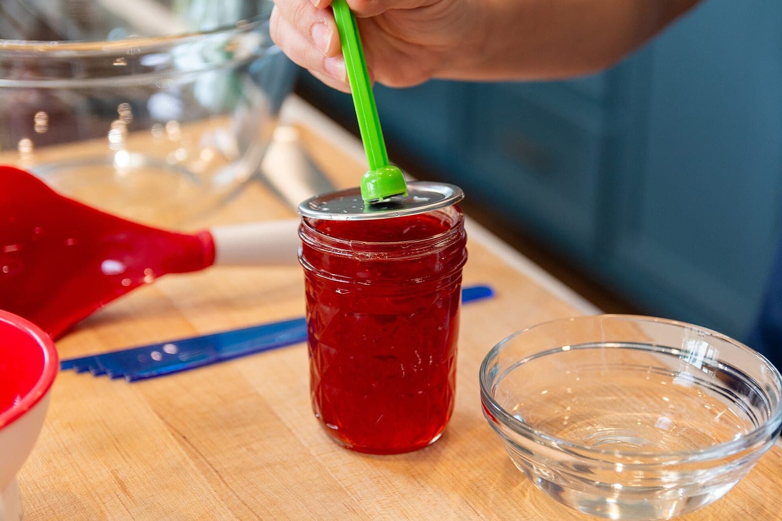 A hand uses a green magnetic wand to set a lid on a jar of red jam beside a small glass bowl