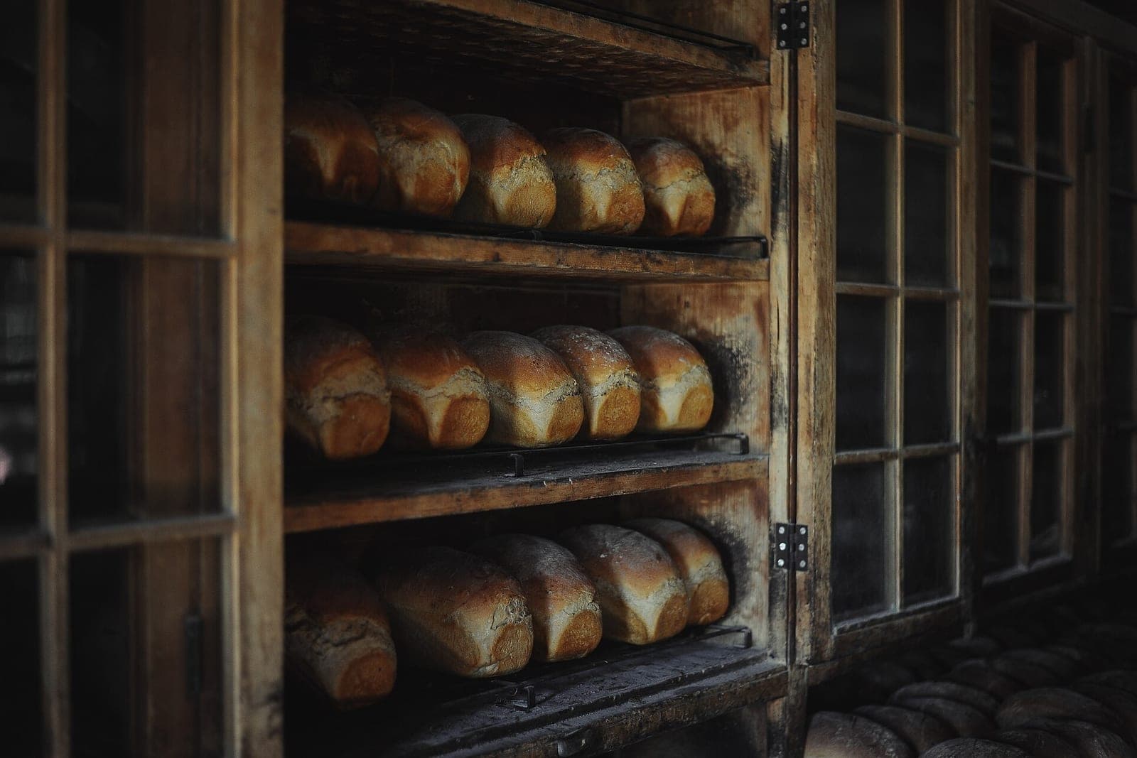Rows of baked loaves of bread on the shelves of a wooden rack with glass doors
