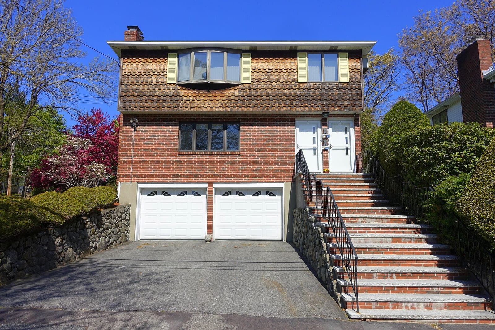 A brick and cedar-shingle house with two white garage doors at the end of a short driveway, beside a tall brick stairway