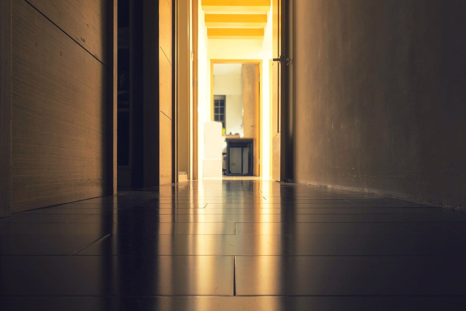 A quiet hallway with a polished floor and an open door to a lit room at the far end