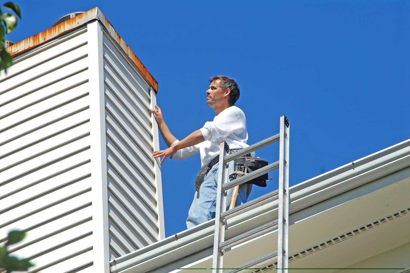 A worker in a tool belt stands at a roof edge beside a ladder and a white gutter