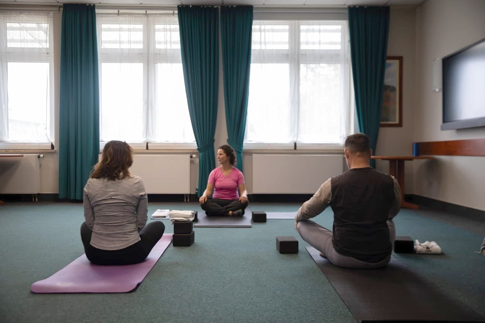 Two members sit on mats facing an instructor at the front of a small exercise class by tall windows