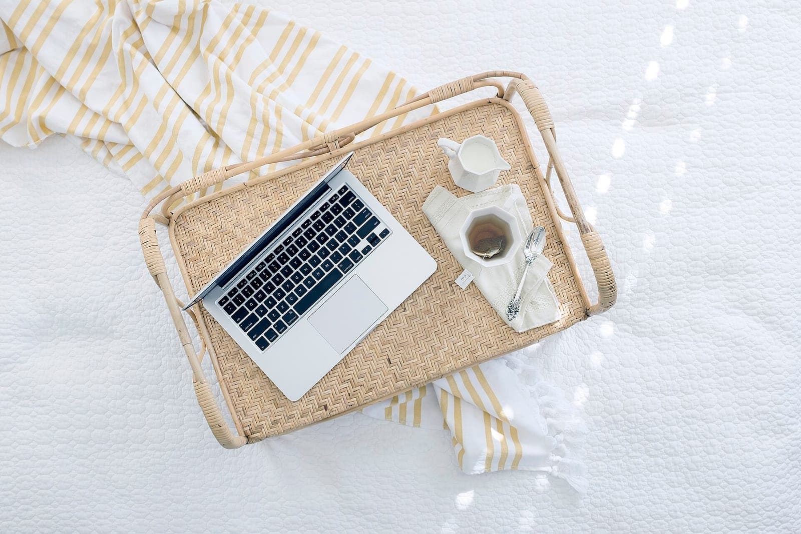 A laptop, a cup of tea and a small milk jug on a wicker tray resting on a white bedspread