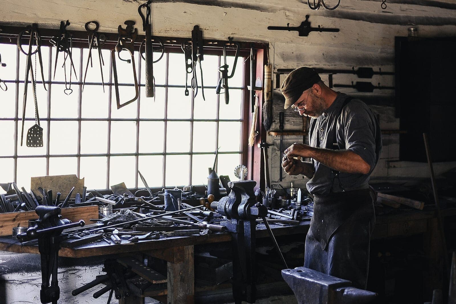 A man in a cap works with his hands at a long workbench by a bright window, with tools hanging above