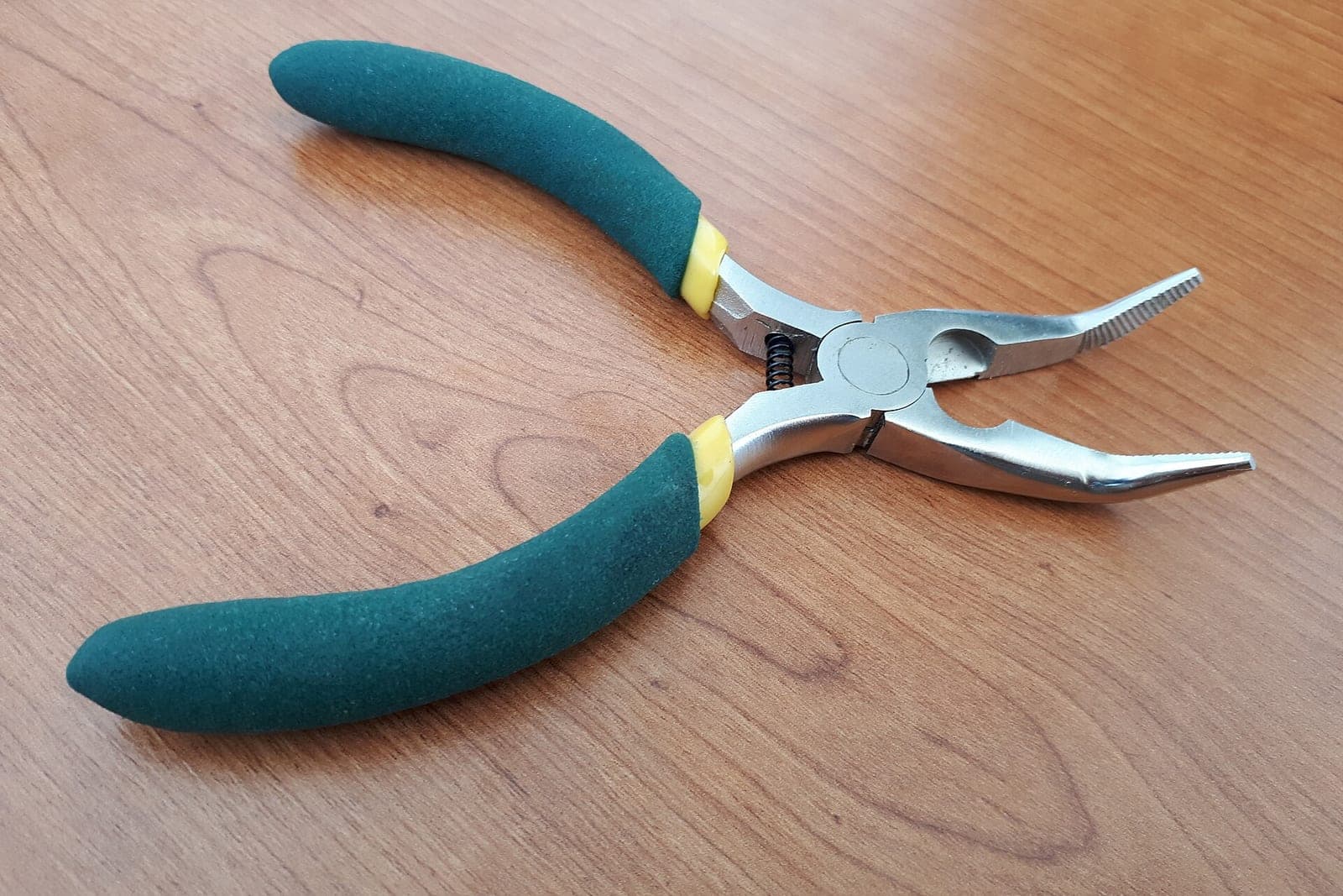 A pair of bent-nose pliers with green grips lying open on a wooden table