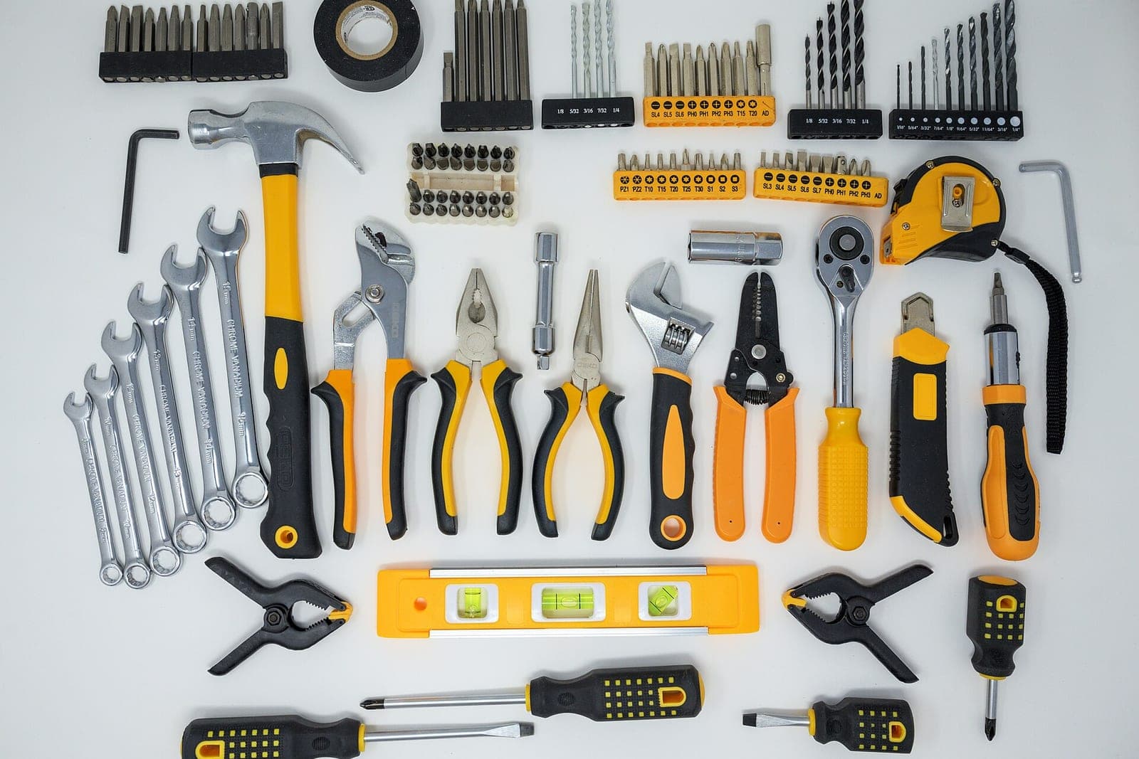 A set of hand tools laid out on a white surface: a hammer, wrenches, pliers, screwdrivers, drill bits, a level and a tape measure