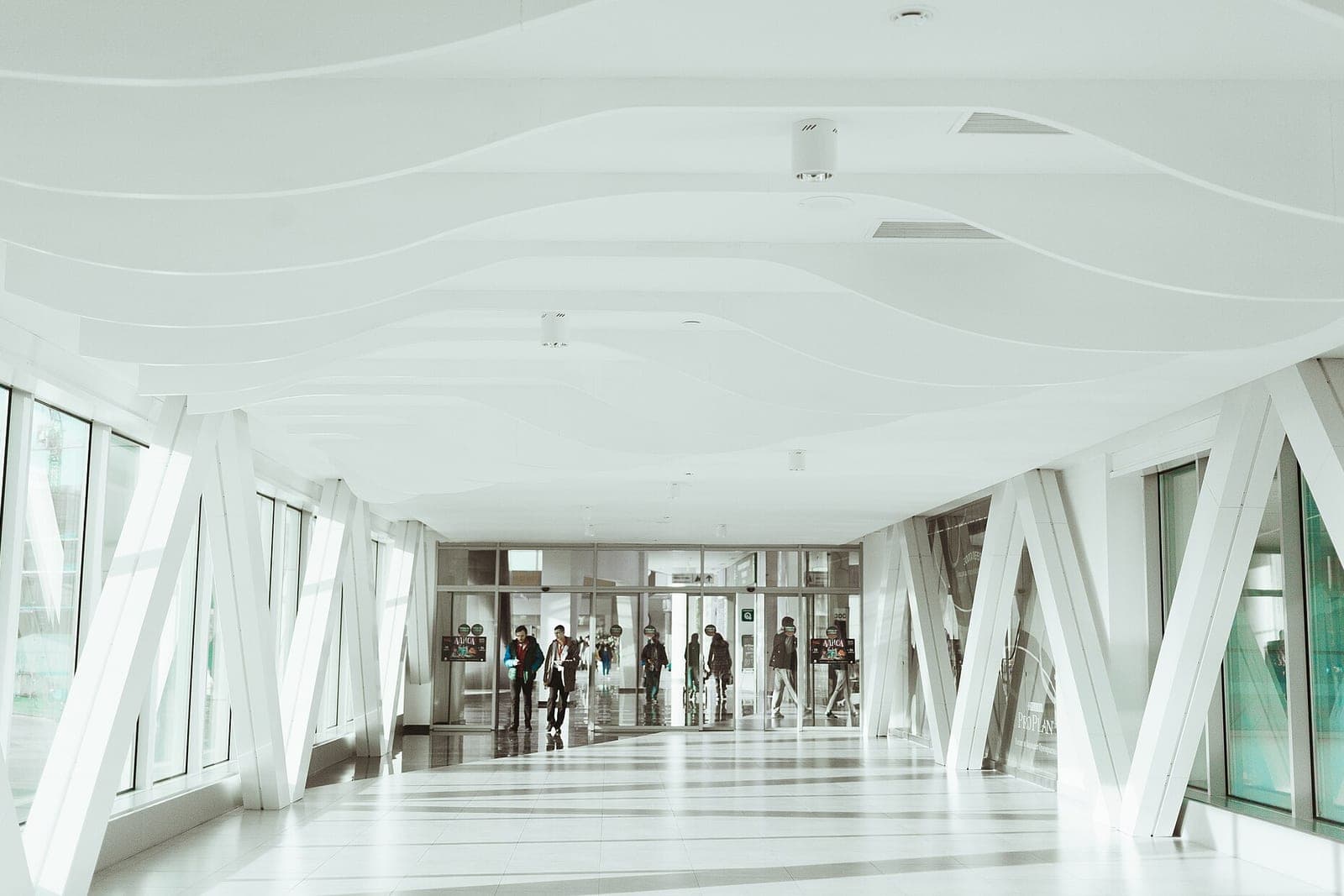 A bright white walkway with angled white beams and glass doors at the far end, with people walking in the distance