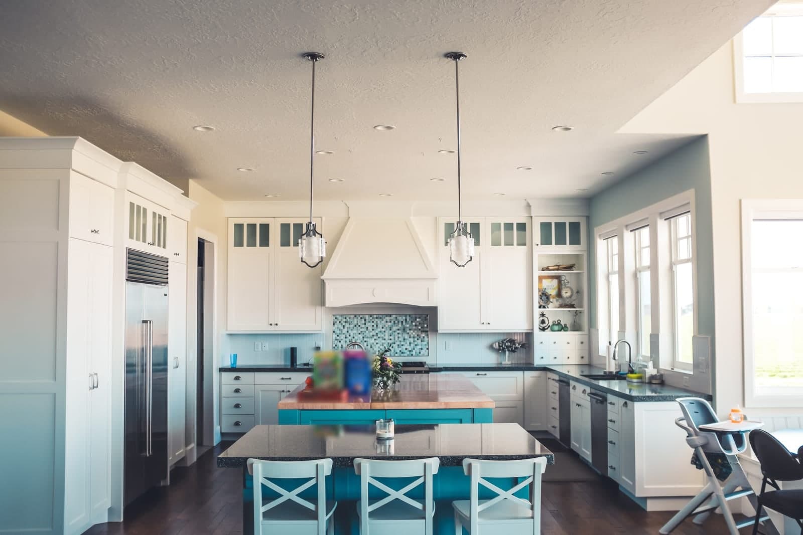 A sunlit home kitchen with white cabinets, a teal island and dark stone counters