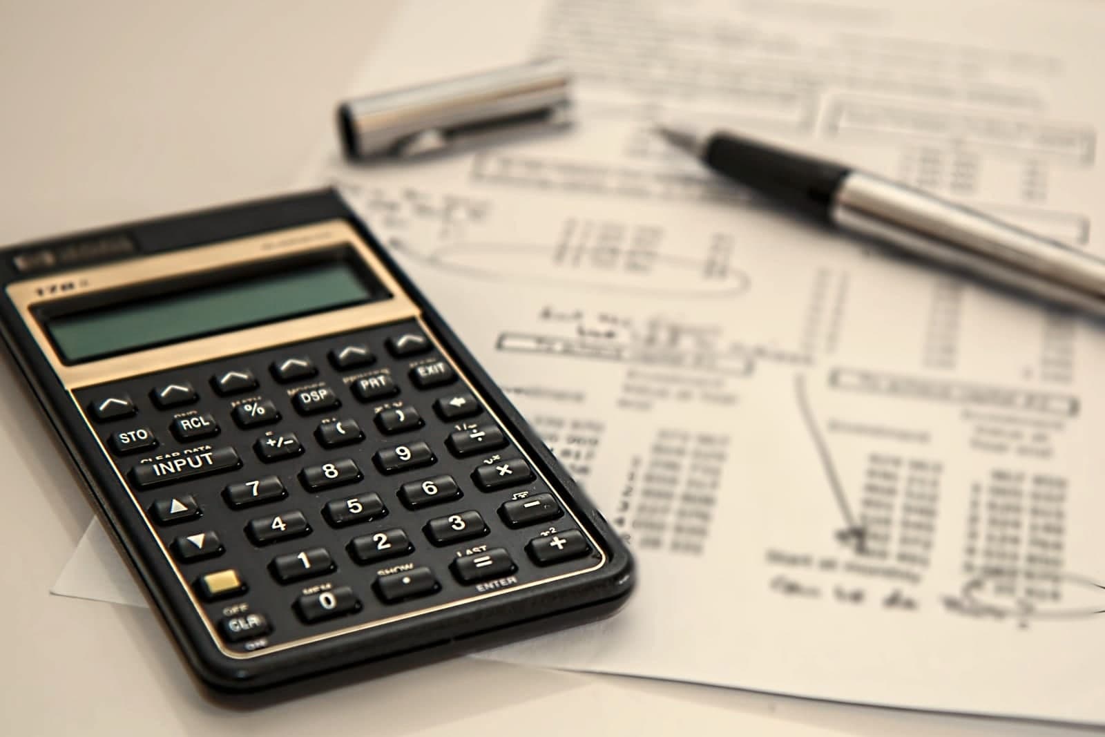 A calculator and a pen rest on printed pages of figures on a white desk