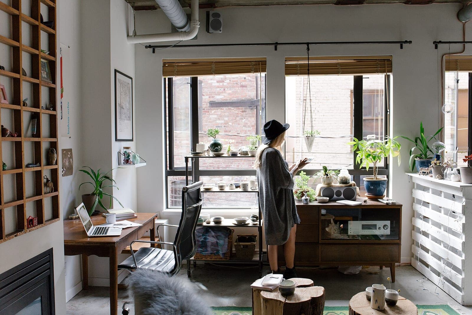 A woman in a black hat stands by the windows of a bright studio office with plants, shelves and a wooden desk