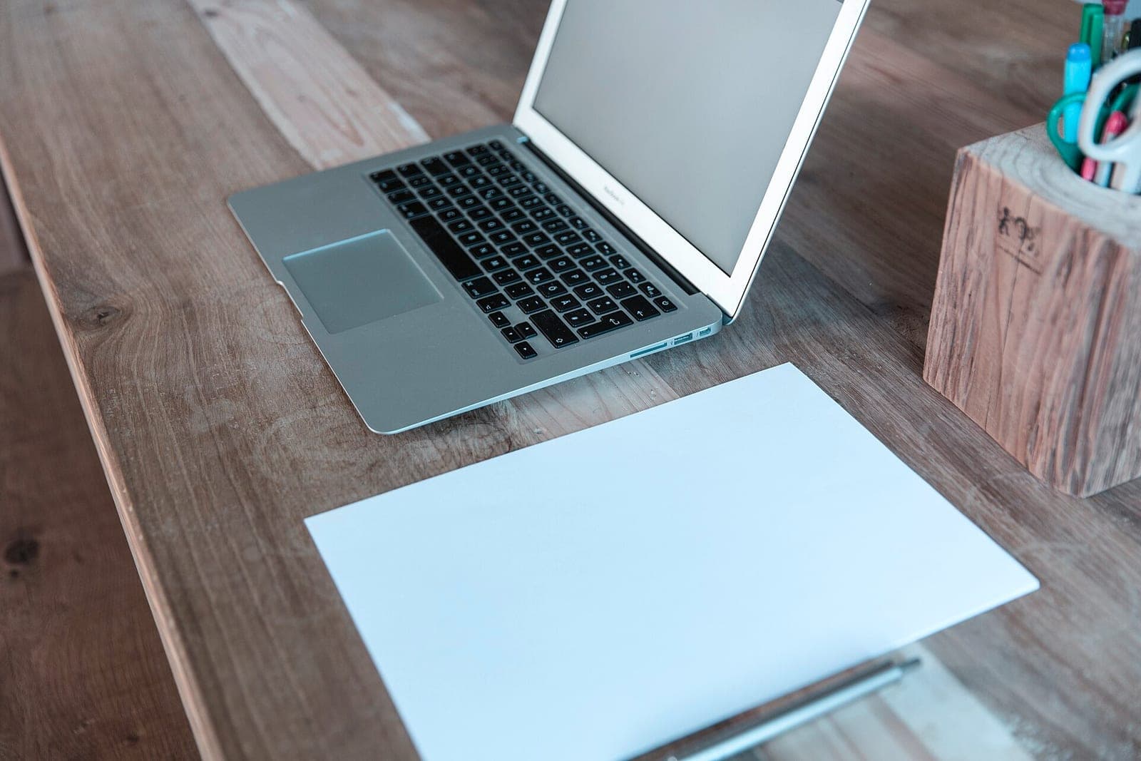 A laptop and a blank sheet of paper on a wooden desk beside a wooden pencil cup