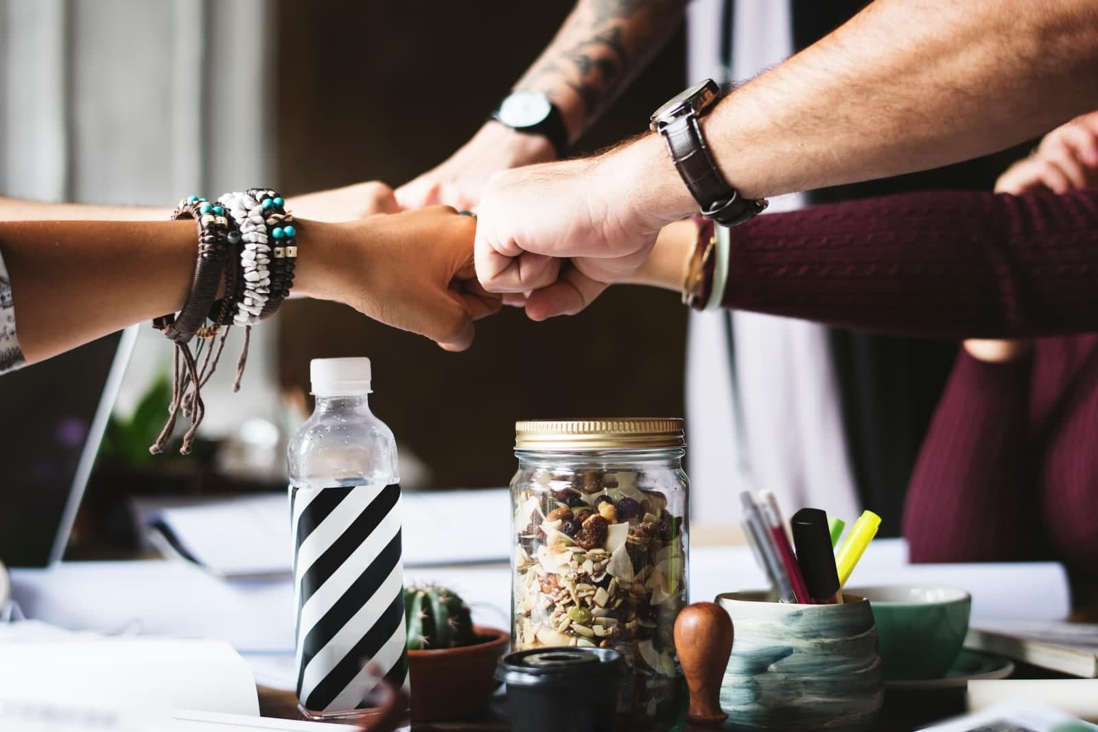 Several hands meet in a fist bump over a work desk with papers, a jar of nuts and a cup of pens