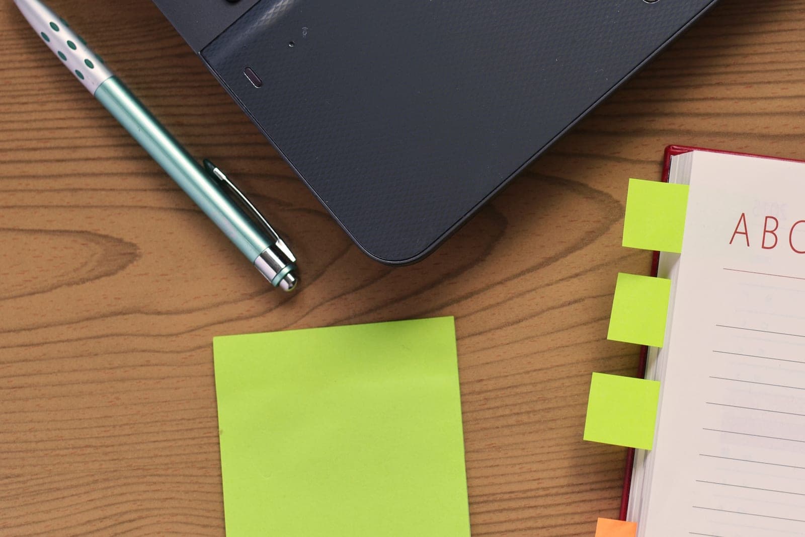 A pen, a blank green sticky note and an open tabbed notebook on a wooden desk