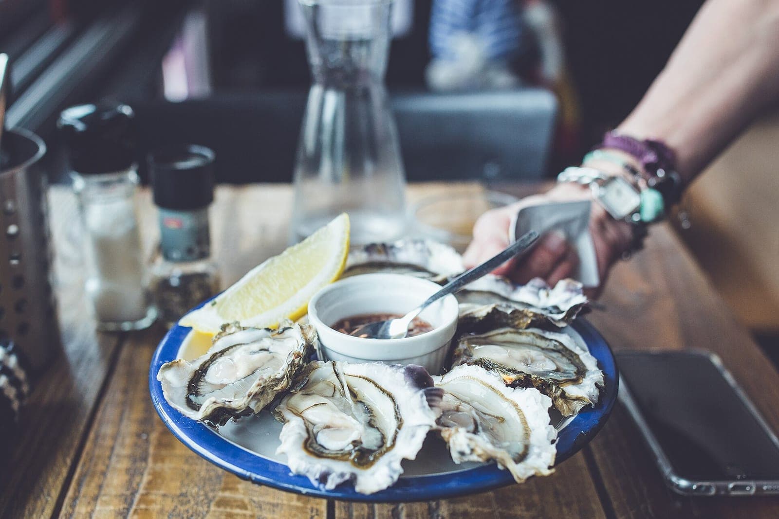 A plate of fresh oysters with lemon and a small dish of sauce being set down on a wooden restaurant table