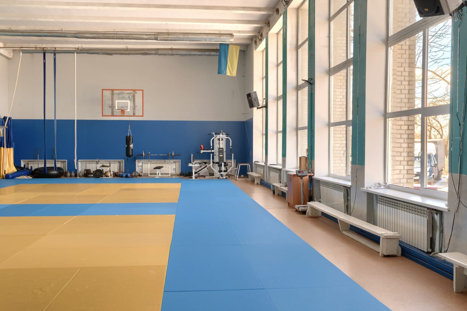 A bright gym hall with floor mats, a punching bag and a weight machine beside tall sunny windows