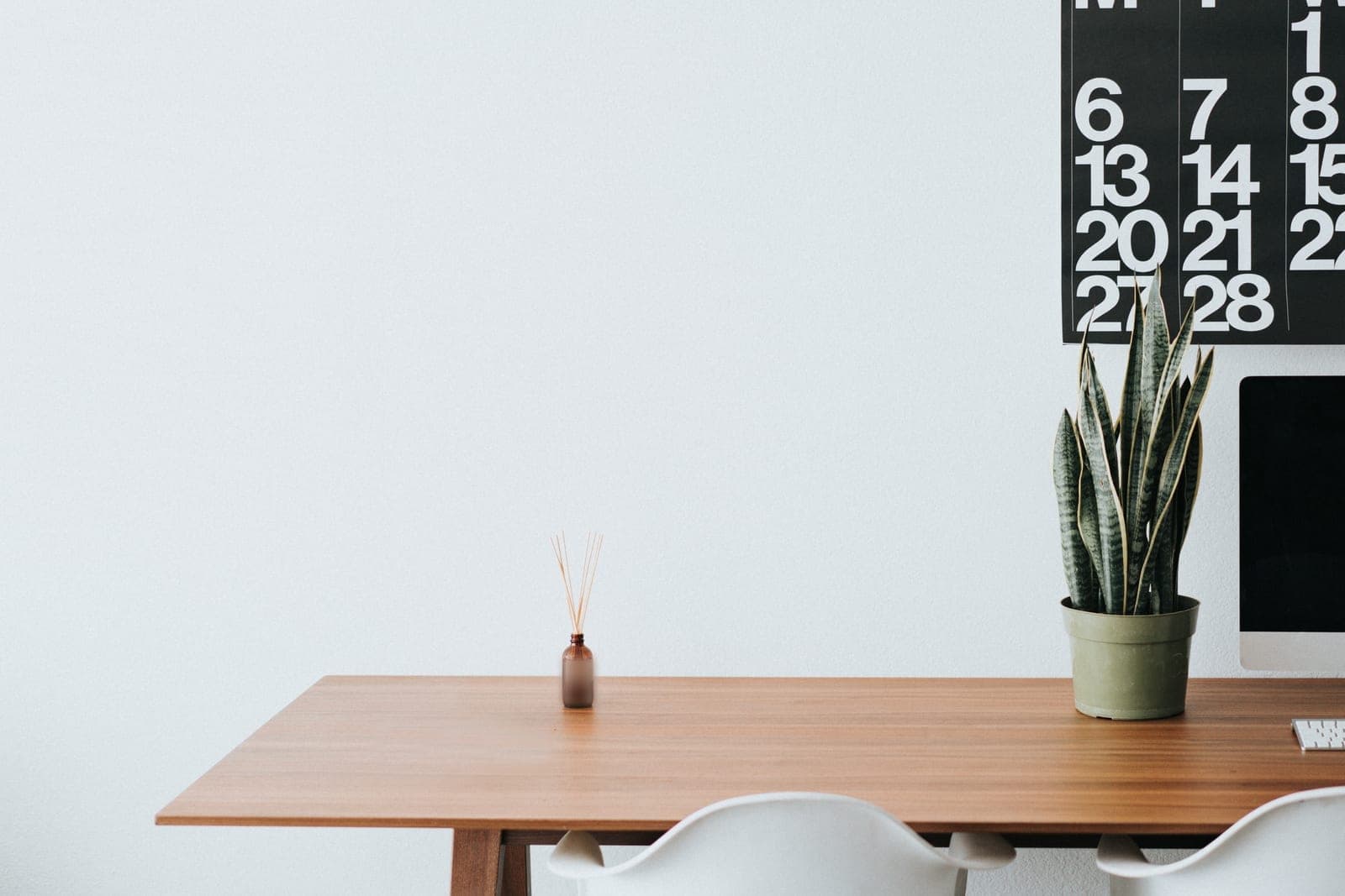 A wooden desk with a potted plant and a monitor below a large black wall calendar