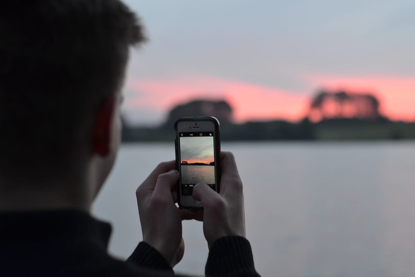 A man seen from behind holds a phone at dusk beside a lake