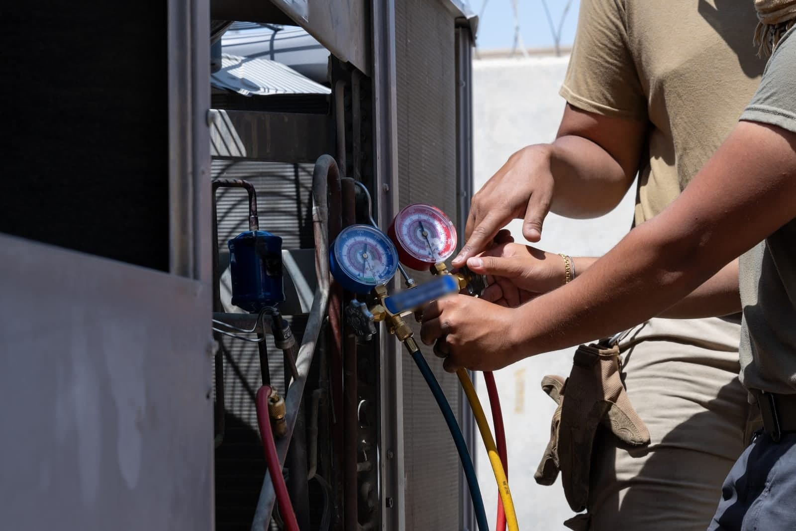 Two technicians' hands connect red and blue pressure gauges to an outdoor air conditioning unit during a service check