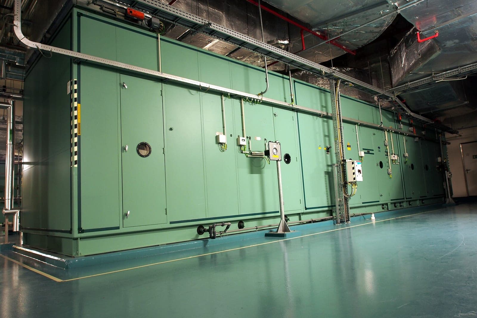 A long green air handler unit with access doors and control boxes in a clean mechanical room under silver ductwork