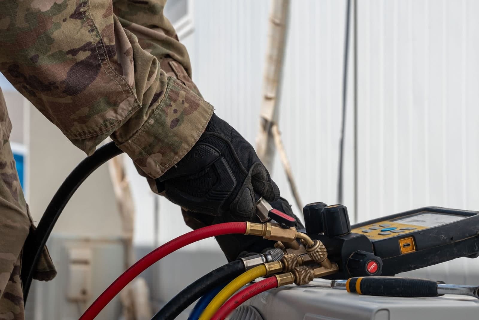 A gloved hand turns a valve on a digital gauge set with red, yellow, blue and black hoses, resting on an air conditioner unit