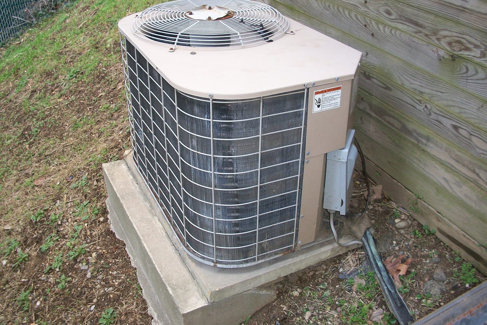 An outdoor air conditioner condenser on a concrete pad beside a wood-sided wall