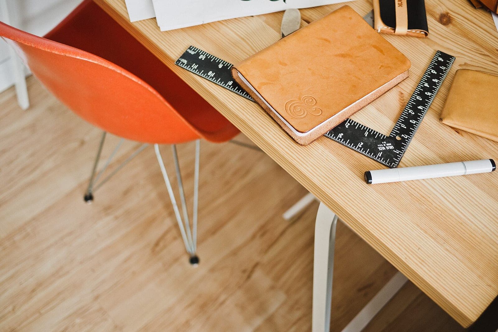 A wooden desk with a leather notebook, a metal ruler and a marker beside an orange chair