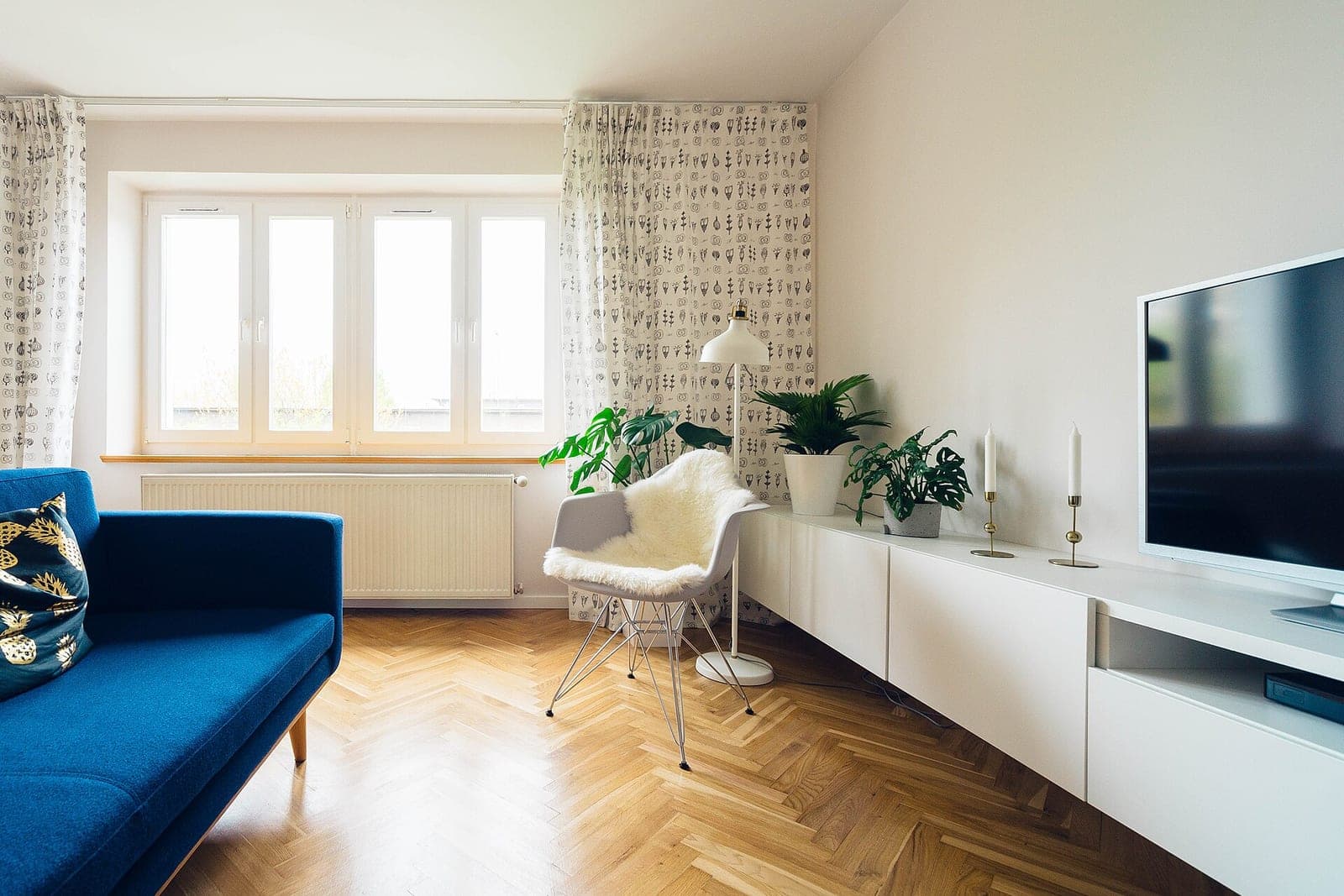 A bright apartment living room with a blue sofa, a white chair, a long white cabinet and a herringbone wood floor