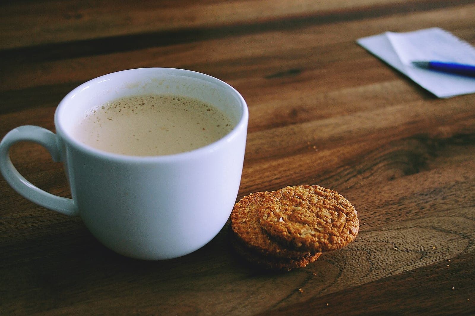 A white mug of coffee and two oat cookies on a wooden table, with a notepad and a blue pen nearby
